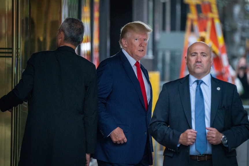 Бывший президент Дональд Трамп выглядит подтянутым в синем костюме и красном галстуке с двумя мужчинами в костюмах по обе стороны.