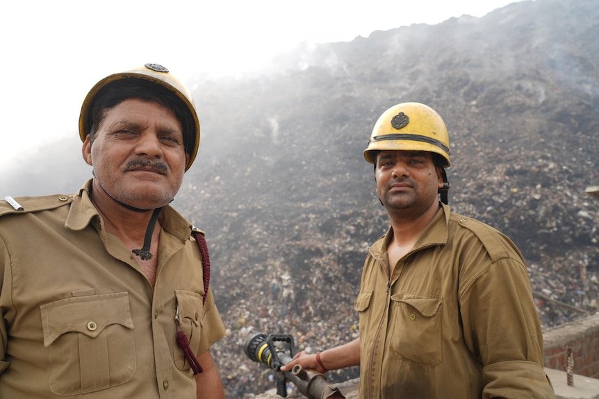 Two Indian men in yellow helmets and brown shirts stand next to a smouldering pile of rubbish