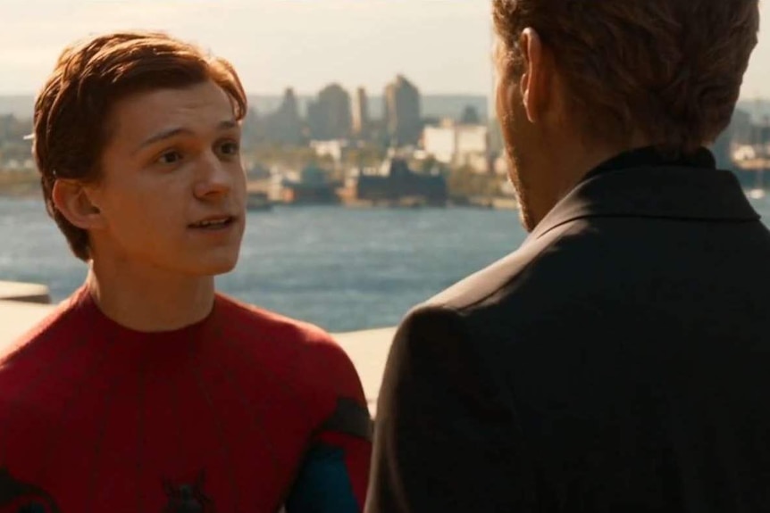A red-haired, shortish white man wearing a Spiderman suit is speaking to another man with his back to camera