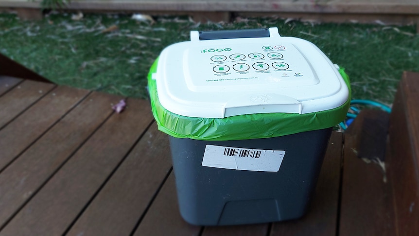 A white kitchen caddy bin with green bin liner sits on a deck outside.
