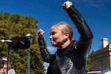 Blonde woman in black jacket stands with fists in air