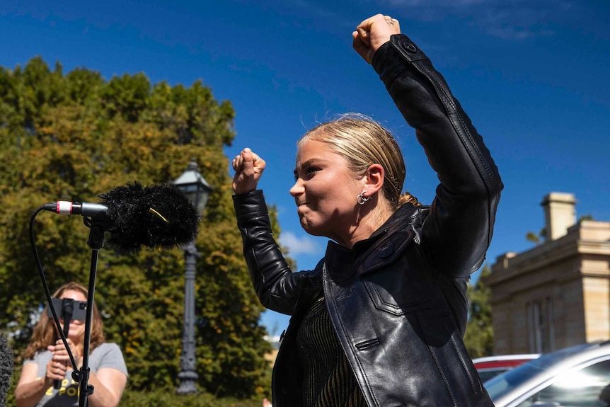 Blonde woman in black jacket stands with fists in air