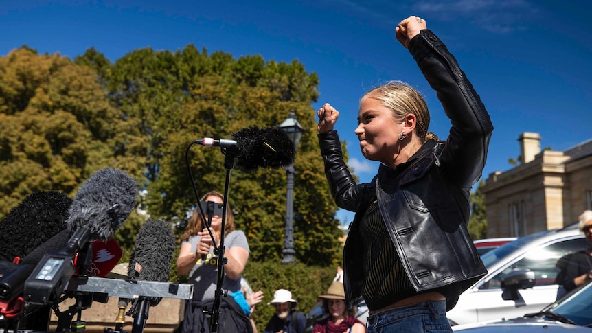 Blonde woman in black jacket stands with fists in air