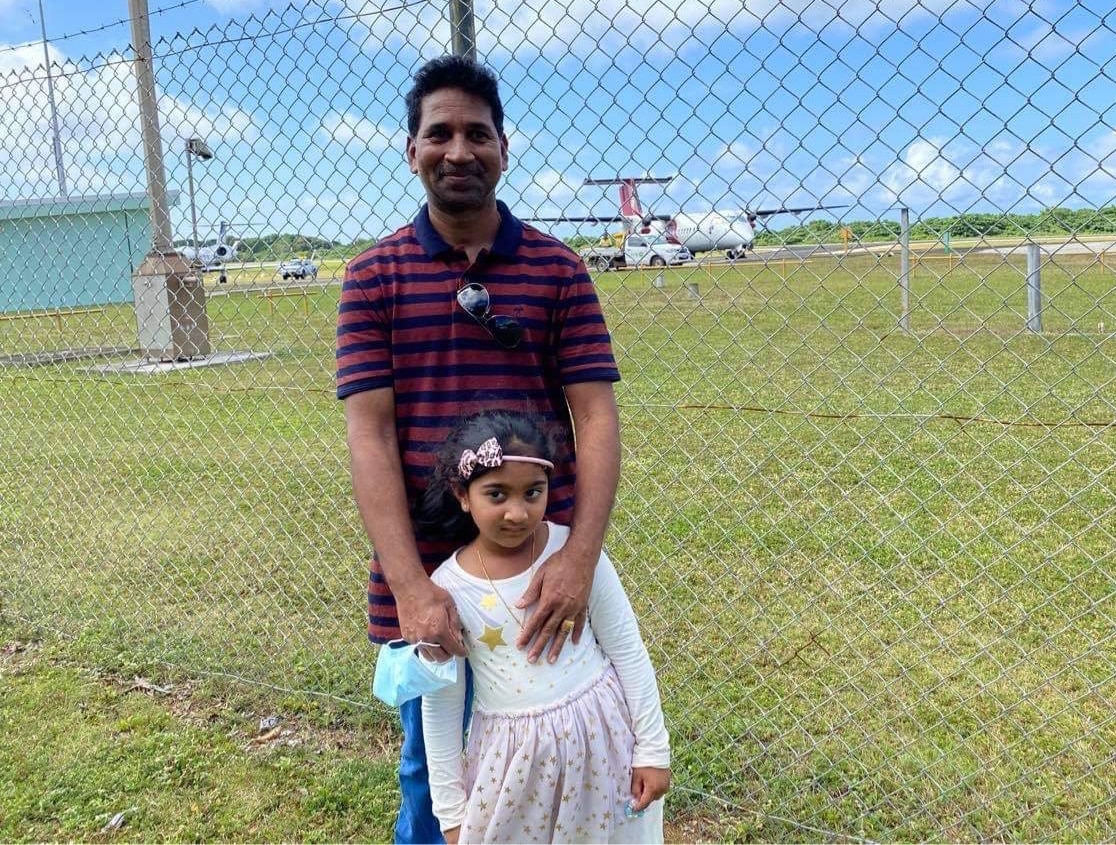 A man smiling as he holds his young daughter in front of an airport fence.