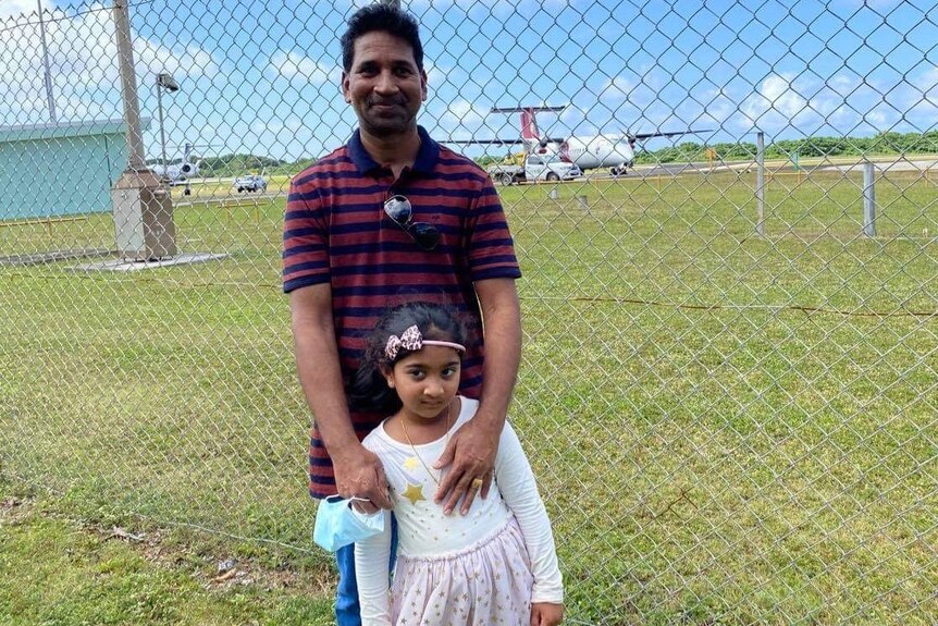 A man smiling as he holds his young daughter in front of an airport fence.