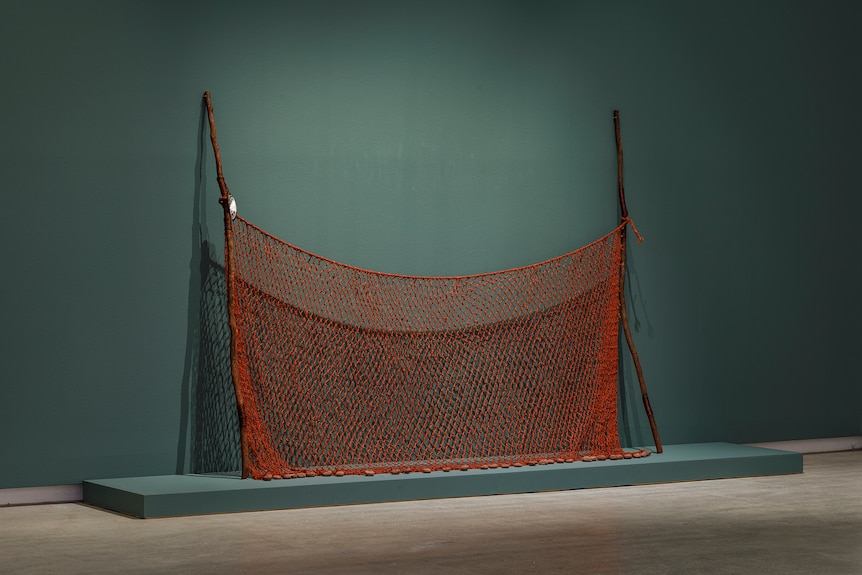 An intricately woven ochre-red net hung across two long dark wooden sticks leaning against a teal gallery wall.