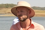 Man in hat under sun talking in front of river.