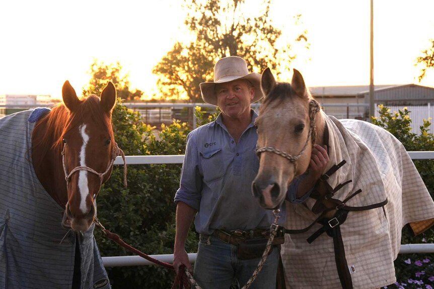 A man standing between two horses with the sun setting behind him