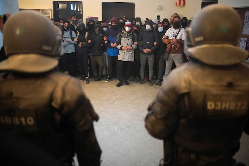 A large group of people linking arms face off against police in helmets and armour.