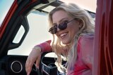 A film still of Renee Rapp smiling brightly behind the wheel of a red carp. She is wearing sunglasses, tilted down on her nose.