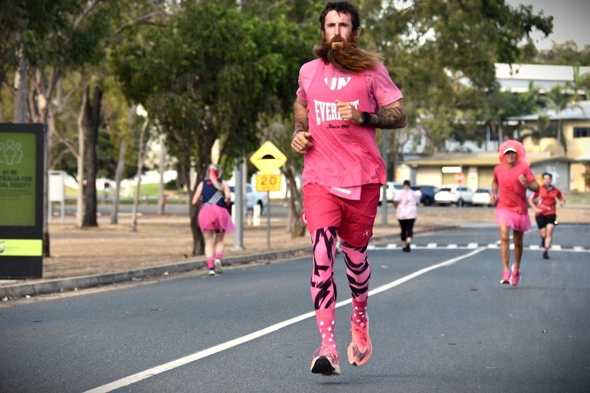 A man with brown hair and brown, ginger beard wearing a pink shirt, shorts and pink and black tights running on the road.