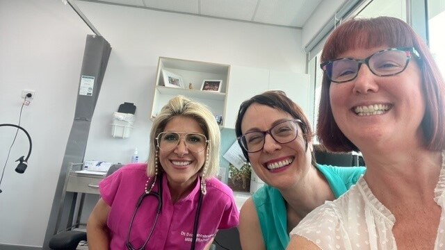 Two women smile alongside a female doctor inside her office.