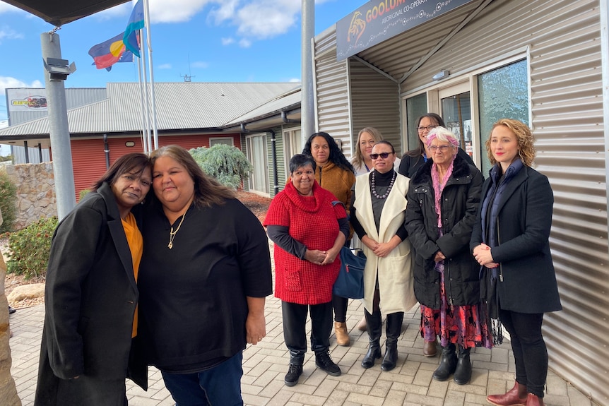 Two Aboriginal woman embrace and pose for a photo in front of a larger group o fwomen behind them. The Aboriginal flag flies in 