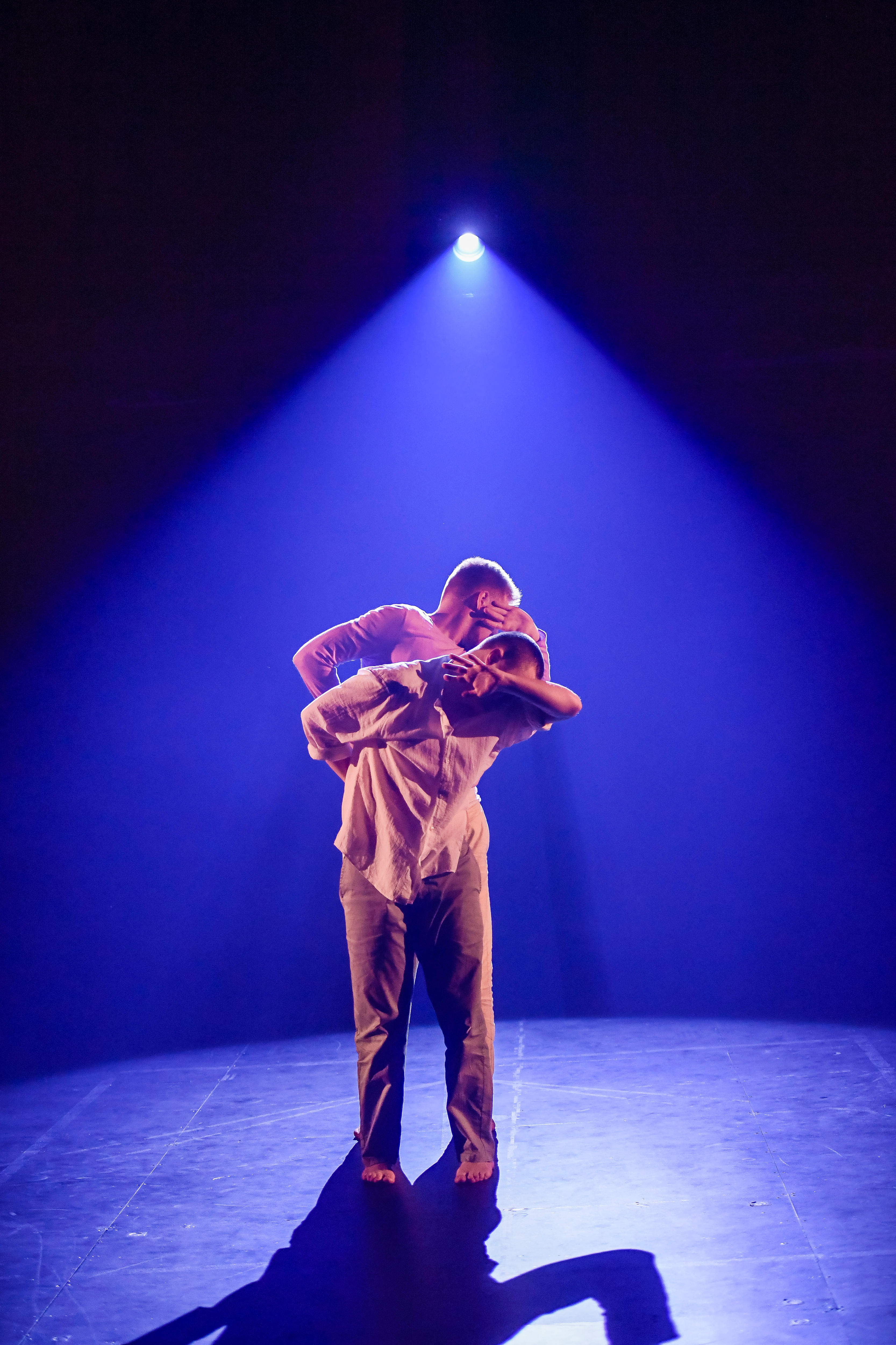 Two white male dancers perform on stage under a sapphire blue light. They shield their faces from the light with their arms.