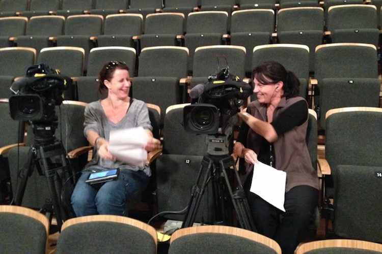 Two women sitting in row of seat with two cameras talking to each other.