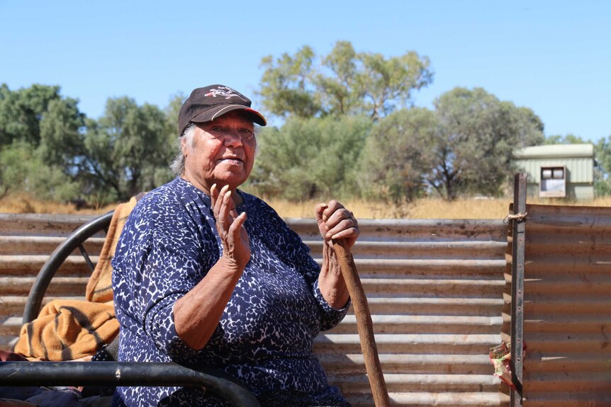 An elderly indigenous woman sits at a camp holding a walking stick
