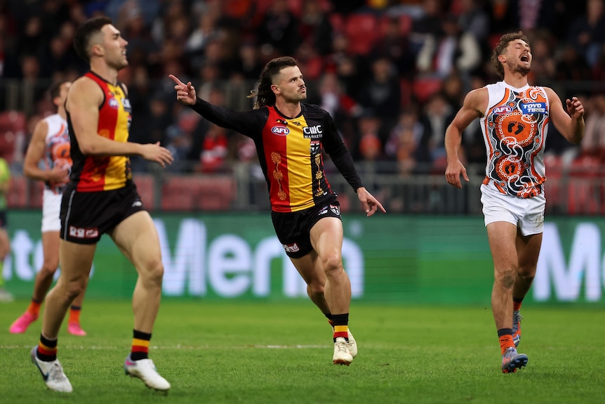 A St Kilda AFL player with a mullet haircut points in celebration as he watches his kick for goal sail through.