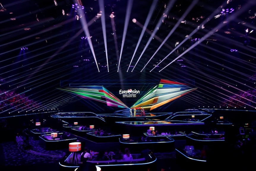 A wide shot of the Eurovision  stadium. Spotlights beam in every direction against deep purple lighting of the room.