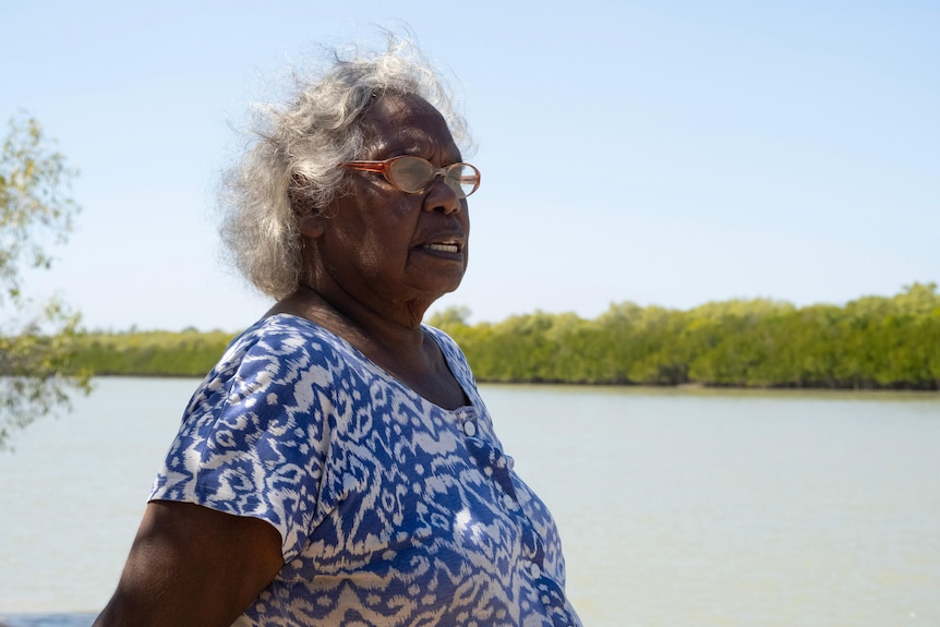 An older Indigenous woman stands by a creek, looking into the distance