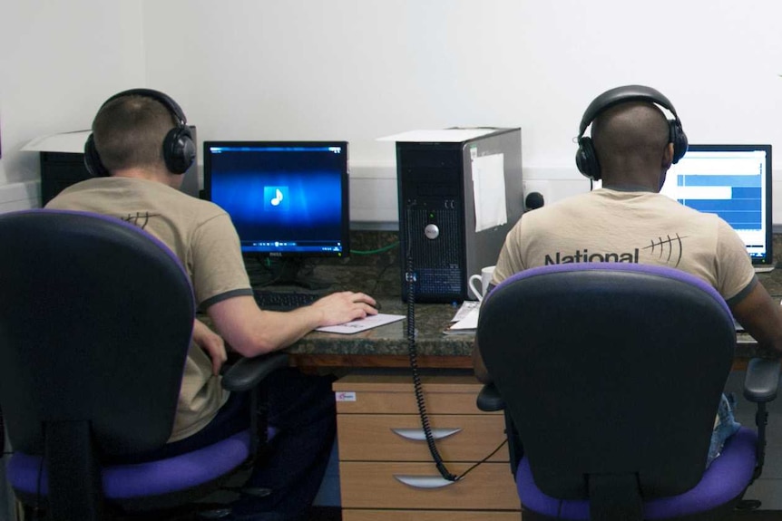 The backs of two men, seated at computers, wearing headphones and wearing National Prison Radio t-shirts.