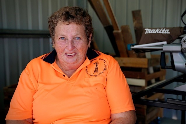 Elderly lady sitting in an open shed wearing an orange high vision shirt.