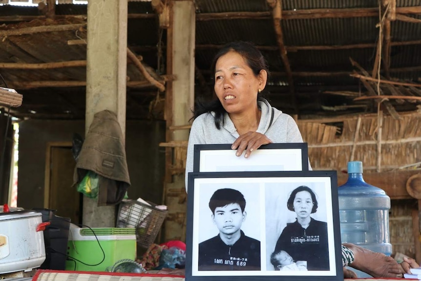 A Cambodian woman sits in a bamboo thatch home holding two black and white mugshot photos.
