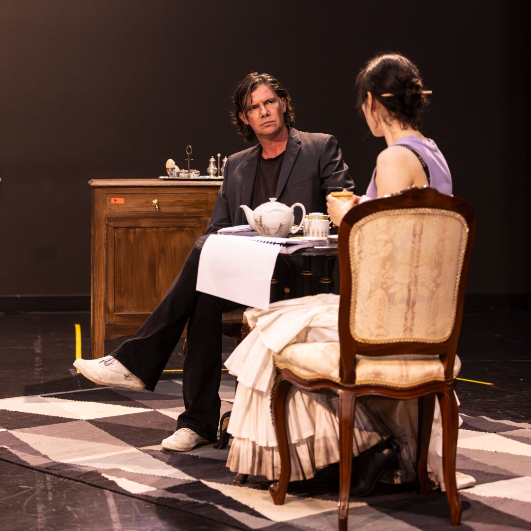 A middle-aged white man looks disapprovingly across a table at a seated 30-something white woman.