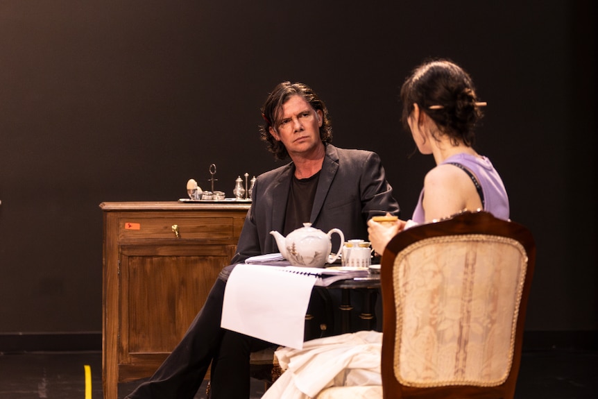 A middle-aged white man looks disapprovingly across a table at a seated 30-something white woman.