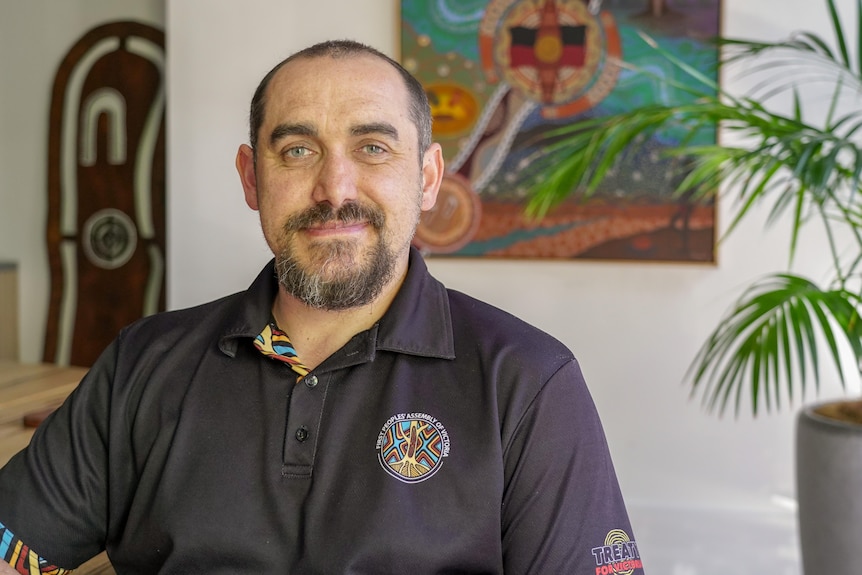 A man in a First Peoples' Assembly shirt, in front of Indigenous art.