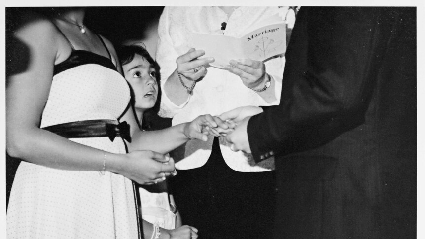 Black and white wedding photo of couple holding hands with young flower girl peering up at the groom in awe