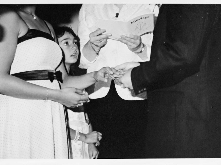 Black and white wedding photo of couple holding hands with young flower girl peering up at the groom in awe