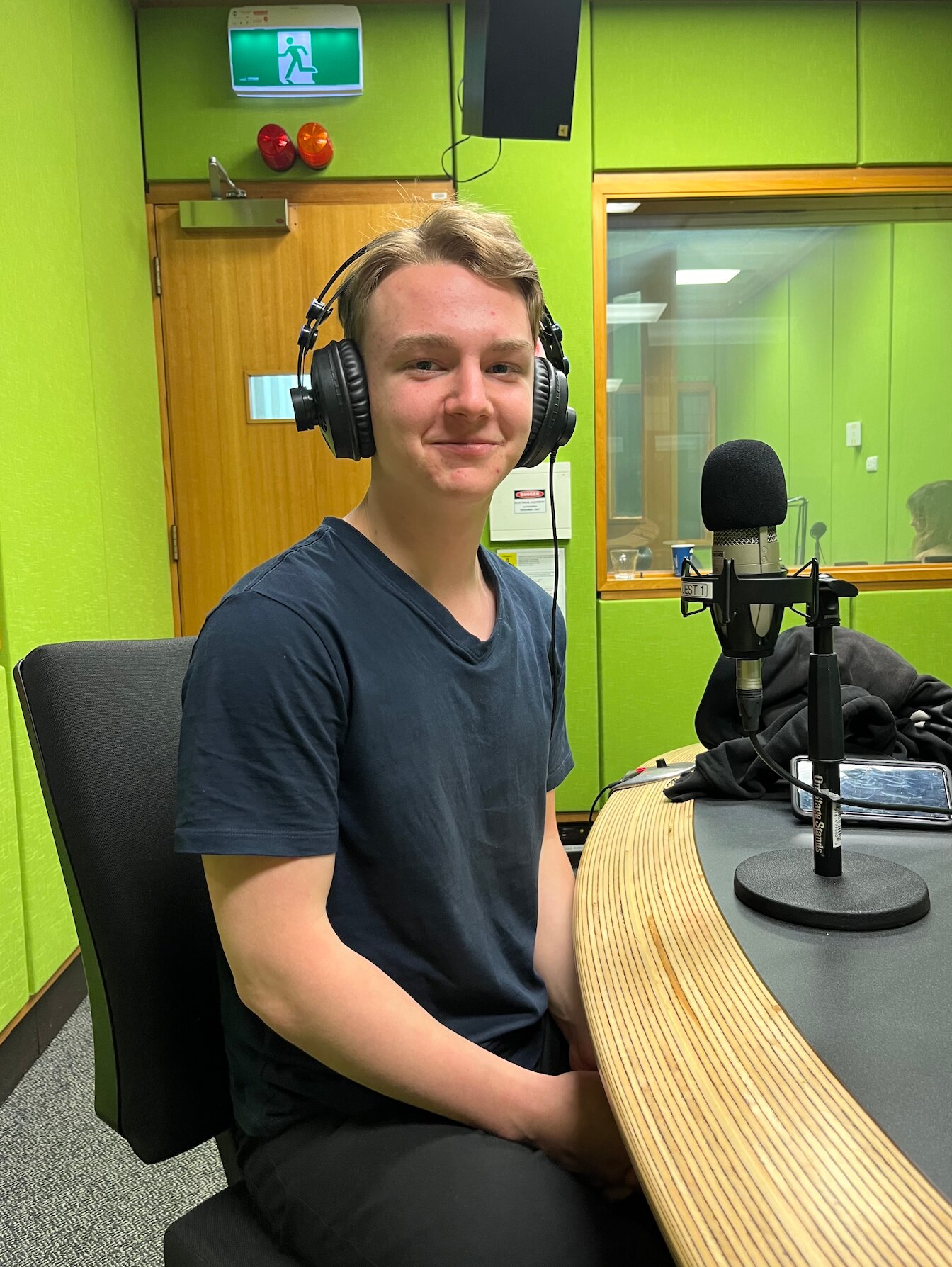 A young man sits in a radio studio with headphones on, smiling at the camera.