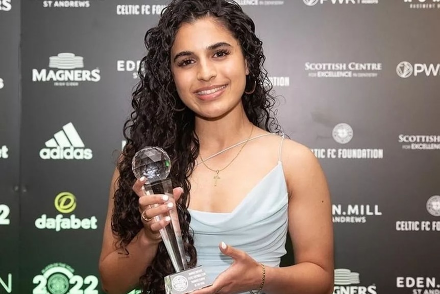 A woman in a grey dress smiles while holding an award in front of sponsor logos