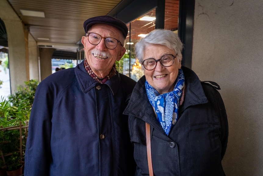 An older couple wearing glasses and in blue jackets pose for a photo while smiling.