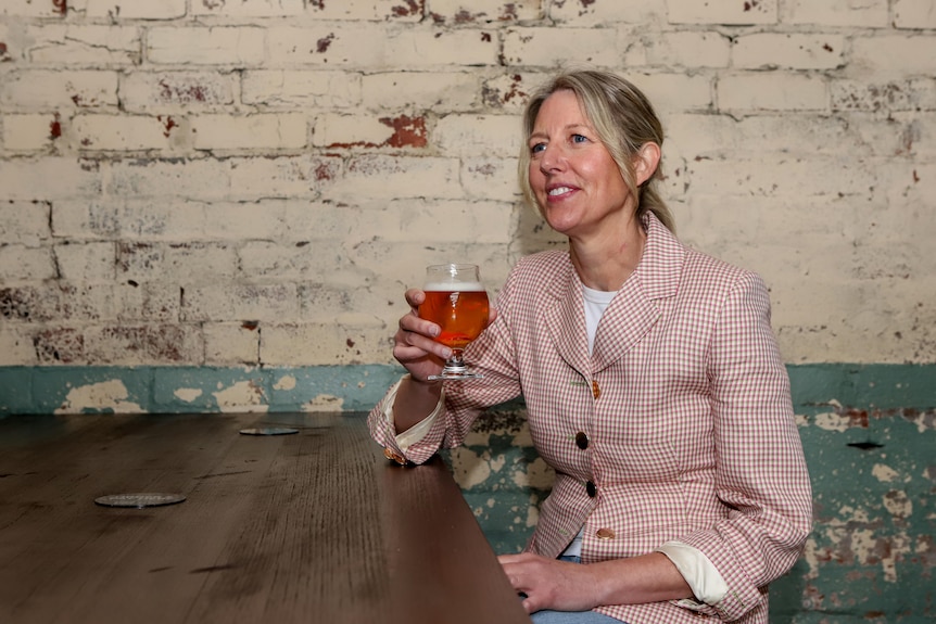 Blonde woman in pink blazer sits at wooden table with glass of beer in hand in front of cracked paint brick wall