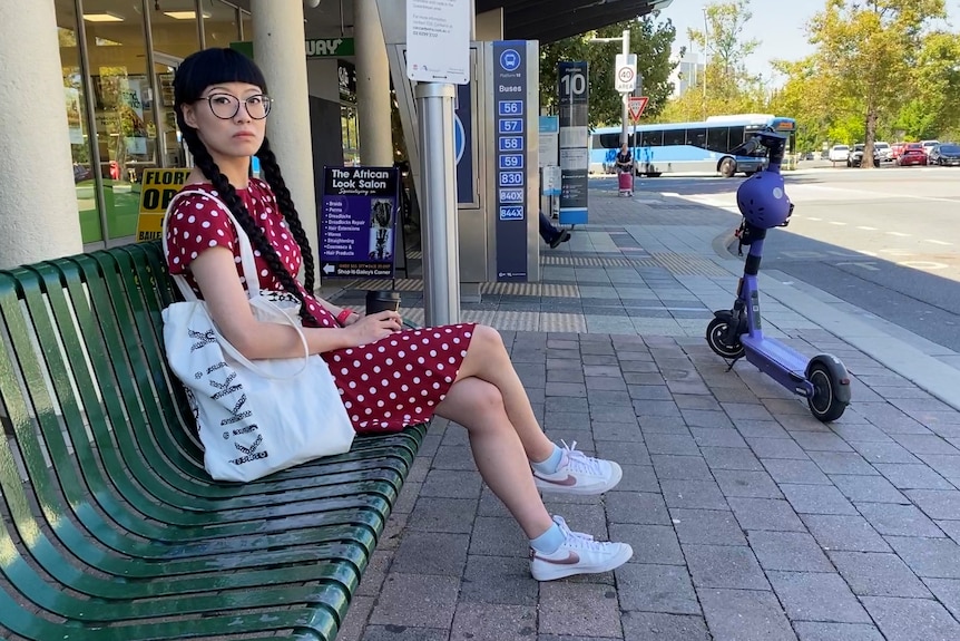 A woman with long black hair sits on a bus bench.