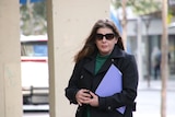 A woman wearing sunglasses and a jacket walks with a folder tucked under her arm.