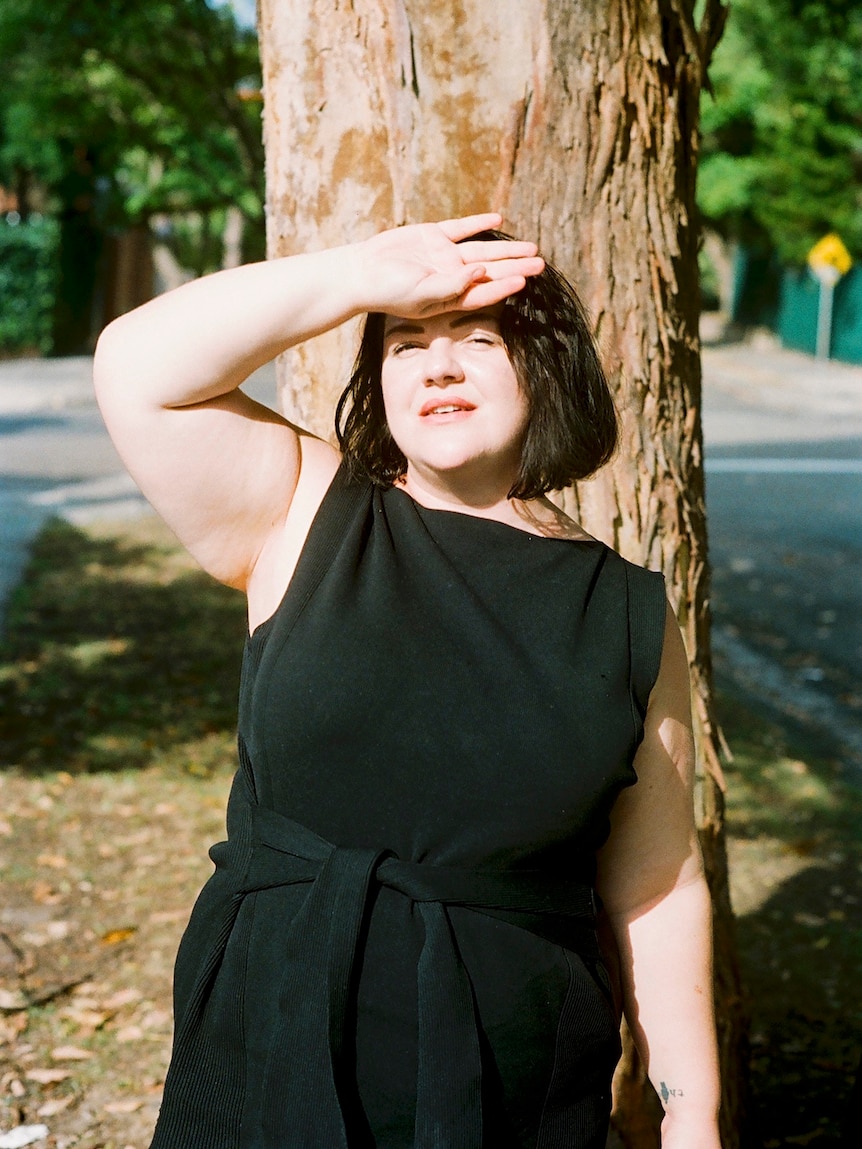 Alison Whittaker stands in front of a tree, with her hand on her forehead to protect her eyes from the sun
