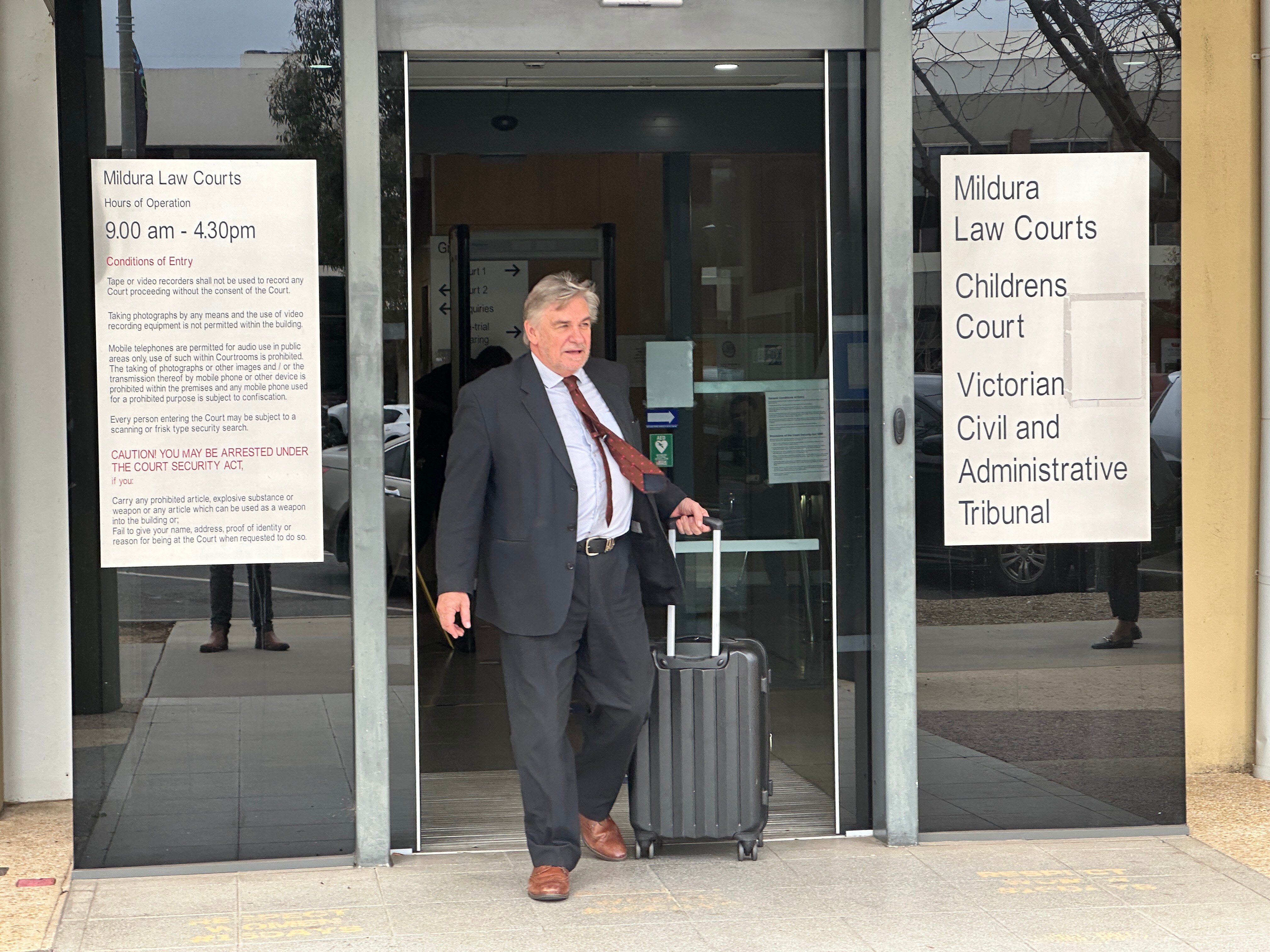 A man in a suit wheels a suitcase out of some sliding doors.