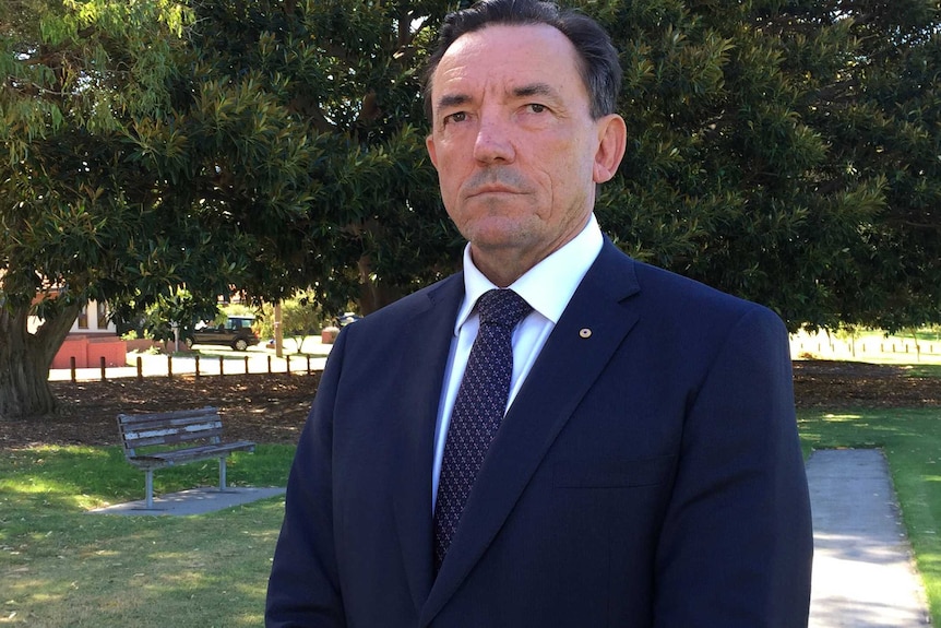 A mid-shot of a stern looking WA Housing Minister Peter Tinley standing outdoors in a park in a suit and tie.