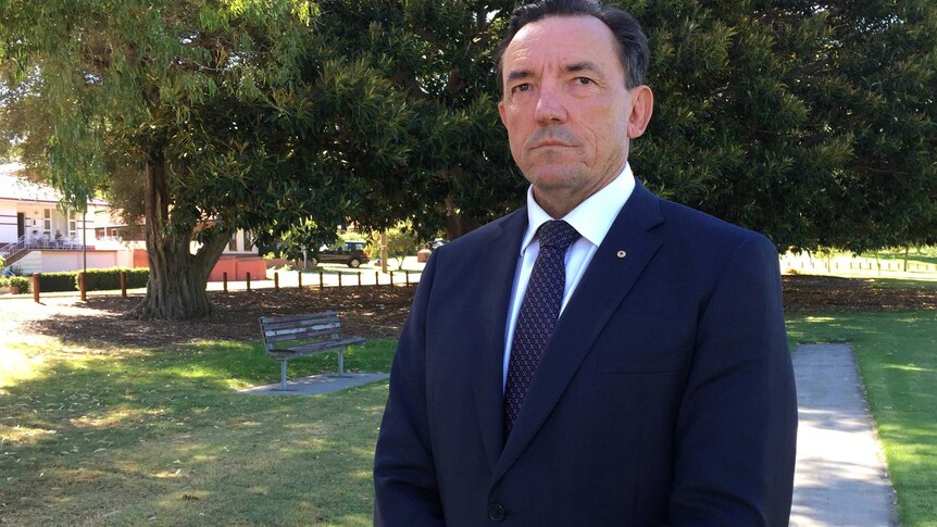 A mid-shot of a stern looking WA Housing Minister Peter Tinley standing outdoors in a park in a suit and tie.