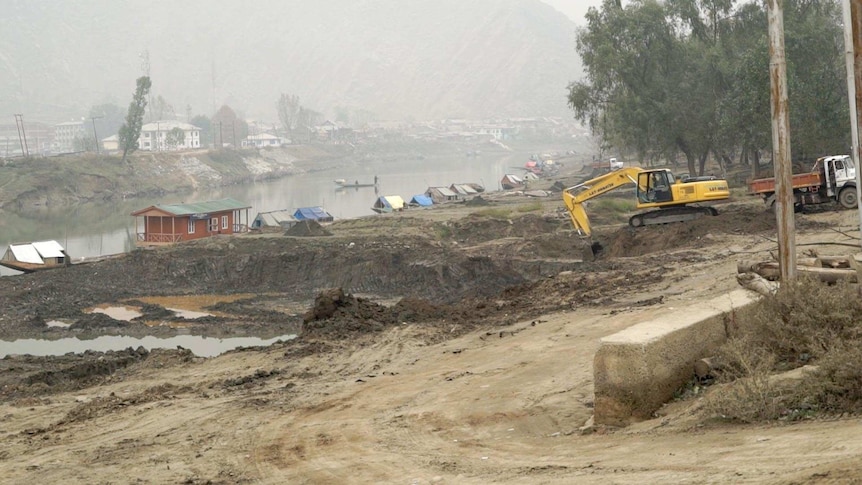 An excavator works on the banks of the river Jellum, with the river and houses across the river in the background.