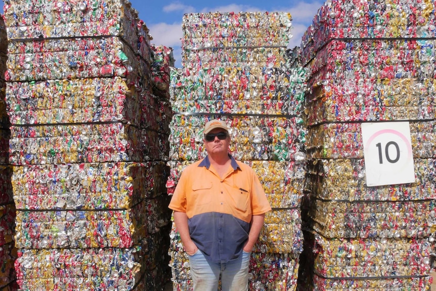 Long shot of Adrian Channing standing in front of bundles of crushed cans piled up high in rows behind him.