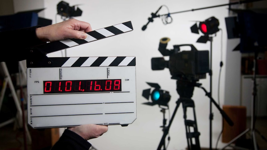 Two hands holding a blank time code film slate in foreground, with cameras, lighting and microphones set up in the background.