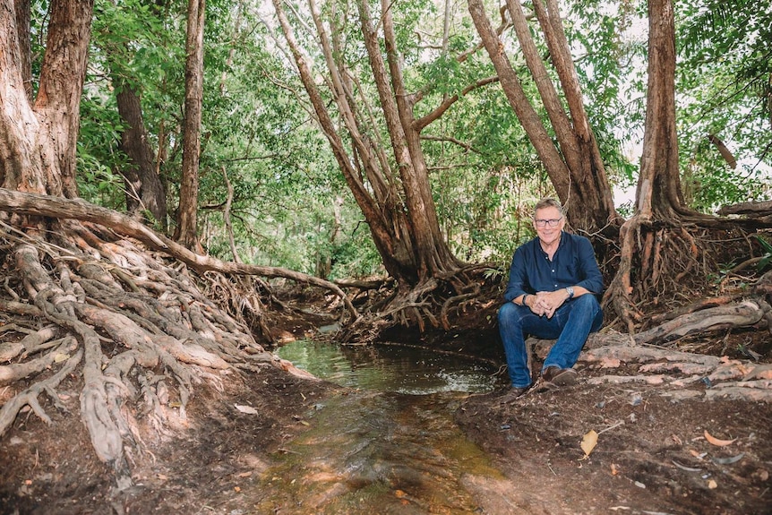 A man in a collared navy shirt and jeans sits on the banks of a creek in mangroves