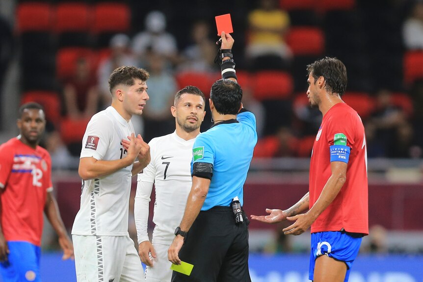 A New Zealand footballer looks up at a referee holding a red card, while his teammate gestures with his hands to his chest.