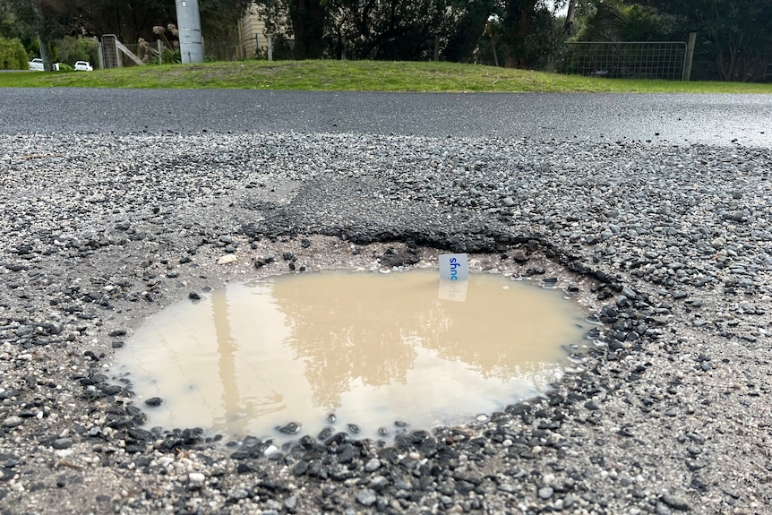 Le premier plan montre un grand nid-de-poule rempli de pluie et un arrière-plan de marqueur montre la route et l'herbe 