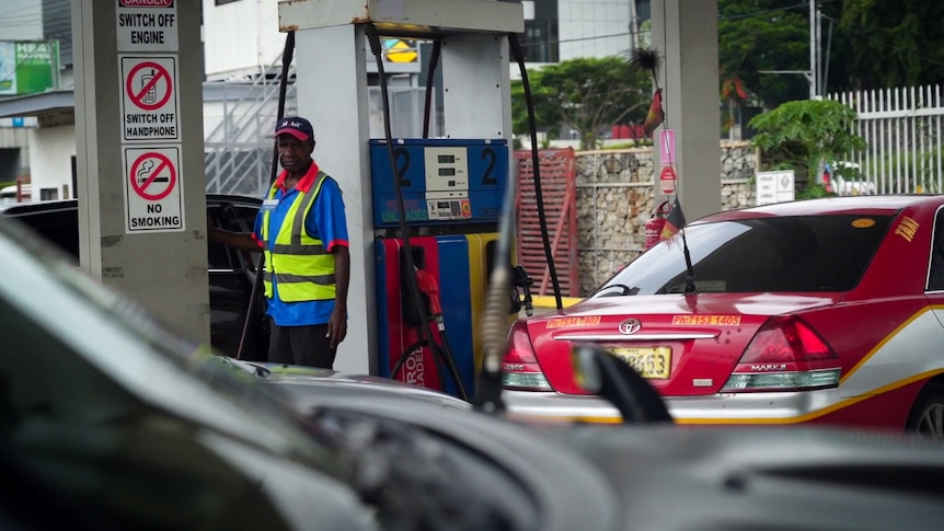 A man wearing hi vis vest over blue shirt with red collar fills a car with fuel, next to a pump