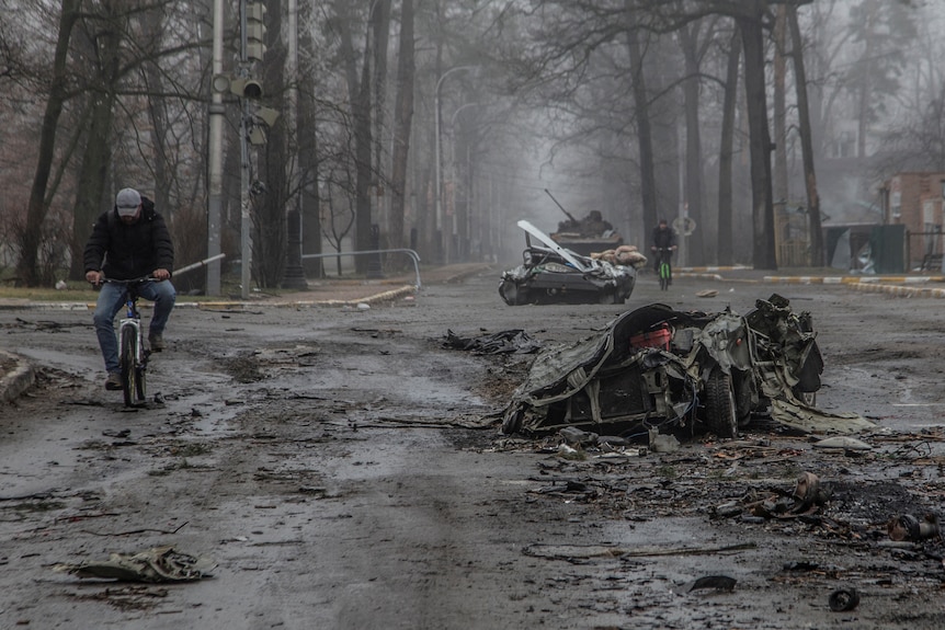 Local residents ride bicycles past flattened civilian cars in Bucha