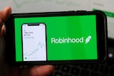 An iPhone is held up to the camera with a green Robinhood ad taking up the entire screen. Behind, a laptop displays stocks chart